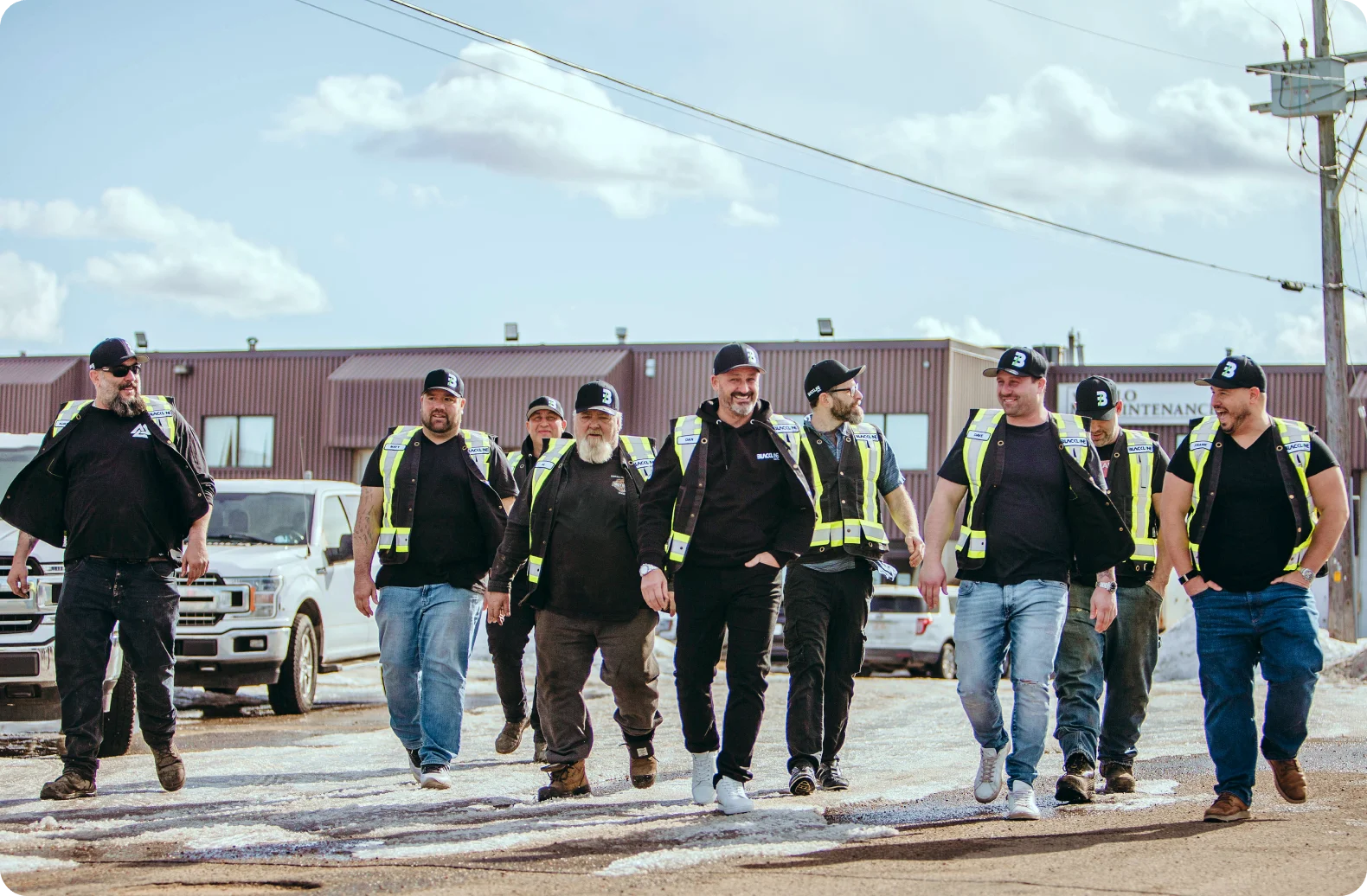 An outdoor, wide shot shows a group of nine men walking confidently toward the camera on a paved area with patches of melting snow. They are dressed in workwear, including black t-shirts or hoodies, jeans, and high-visibility safety vests. Many are wearing black baseball caps with a white 'B' logo. In the background, there is a brown industrial building, a white pickup truck, and a clear blue sky with light clouds. The group appears to be a professional crew or team, captured in a candid, 'hero walk' style.